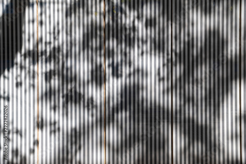Abstract pattern of soft tree shadows falling across corrugated metal wall, creating alternating vertical stripes of sunlight and shade.