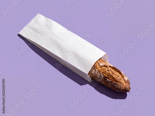 Fresh crusty baguette in white paper bag on purple background