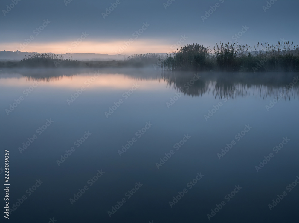Obraz premium Distant reeds and thin fog over still blue water at pre-dawn with clean space below