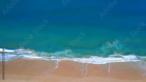 Aerial slow motion tracking view of ocean waves rolling and crashing onto sandy beach shoreline from above