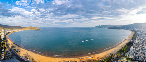 Daytime aerial panoramic drone view of Quy Nhon city coastline, Vietnam. High-angle shot showing a sandy beach crowded with people, crystal clear turquoise sea with boats, long horizon, and a bright