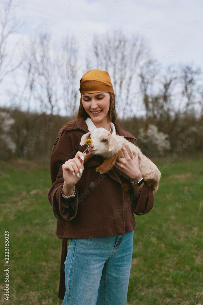 Fototapeta premium A smiling woman outdoors gently offers a yellow flower to a playful goat kid, capturing a warm lifestyle moment in a rural field bathed in natural light and calm atmosphere.