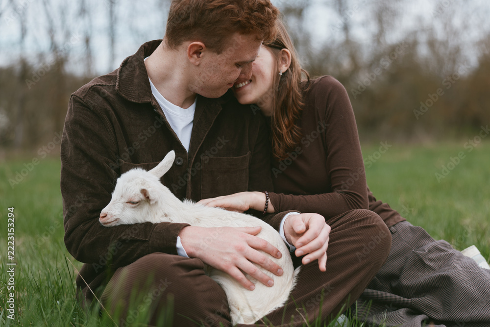 Fototapeta premium A couple sits in a grassy field outdoors, warmly embracing a small goat kid resting on their laps, with tender contact and bright smiles conveying simple lifestyle joys and animal companionship.