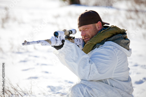 Man in white winter camouflage aims a sniper rifle in snow