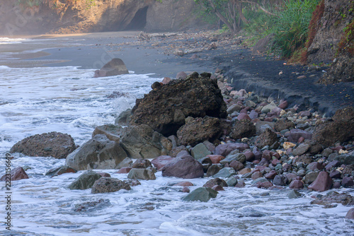 Rugged rocks meet crashing waves in wild coastal scene. Foamy surf rushes over stones in constant motion. Nature's power shapes coastline with raw, untamed force. Ideal for travel, adventure