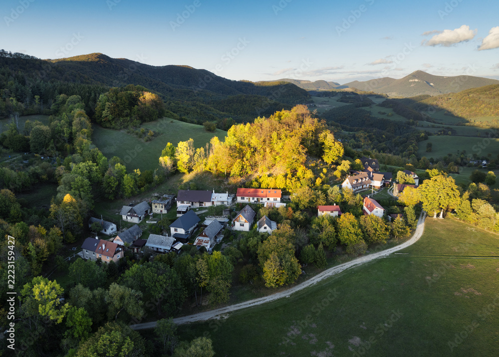 Fototapeta premium Drone view of small mountain village in Slovakia surrounded by forests, and rolling hills in vibrant fall colors, bathed in golden afternoon light.