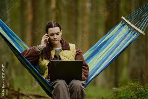Young adult Caucasian woman sitting in hammock in forest, talking on smartphone while working on laptop. Visual representing remote work stress suitable for telecommuting campaigns