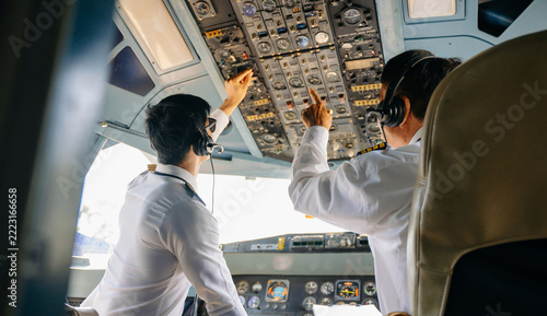Two airline pilots adjust switches on the overhead control panel inside an airplane cockpit during a preflight check.
