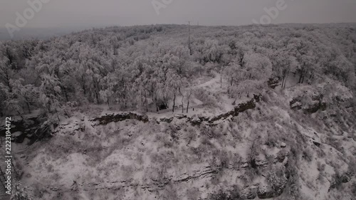 From above, snowy trees fill remote woodland beside rocky cliff. Footpath with wooden railing curves through winter powder beneath gray, overcast sky.