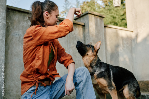 Obraz na plátně A young woman in an orange shirt and jeans trains her german shepherd dog, offering a treat outdoors in the backyard on a sunny day