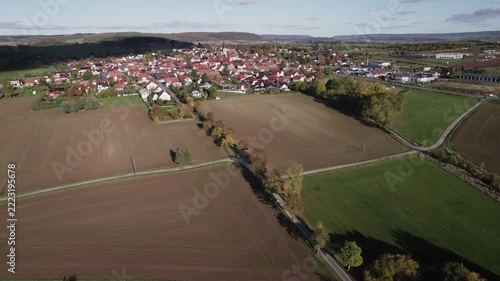 High angle aerial photograph showing compact European town centered on church steeple, red roof houses, autumn foliage, winding streets, and surrounding fields with clear daylight and scenic rural