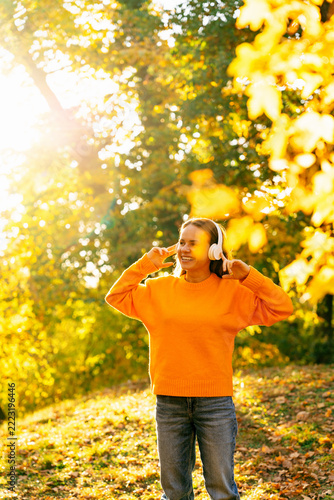 Carefree young woman in headphones enjoying cozy walk in sunny autumn park