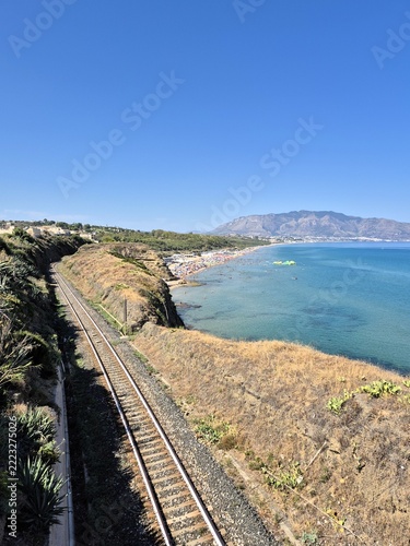  Railroad tracks following a spectacular coastline with panoramic sea views and distant mountains. Transportation and travel concept immersed in a Mediterranean environment.

