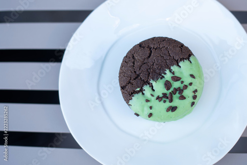 A round chocolate mint cookie with a cracked surface, half covered in green mint icing and topped with chocolate chips, placed on a white plate against a striped background. copy space