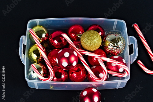 A clear storage bin holds shiny red and gold ornaments, glittery accents, and striped candy canes against a dark background for holiday decorating.