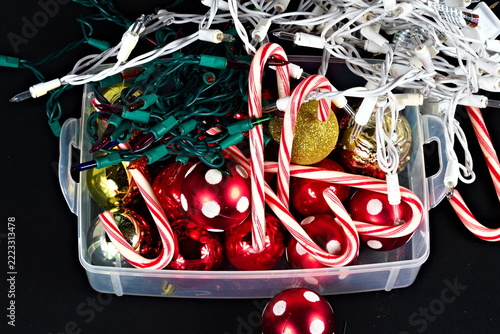 Red ornaments, striped candy canes, glittering gold decorations and tangled white and green Christmas lights fill a transparent container on a dark surface.