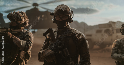 Masked Special Operations Soldier in Ballistic Helmet, Goggles and Tactical Gloves Shoulders His Weapon While His Squad Secures a Dusty Desert Landing Zone by a Helicopter and Armored Truck.