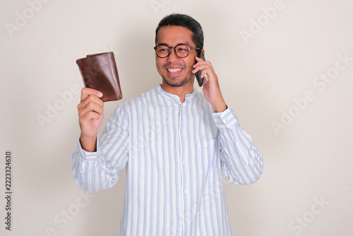 Asian man looking at a wallet that he hold while calling using mobile phone with smiling face