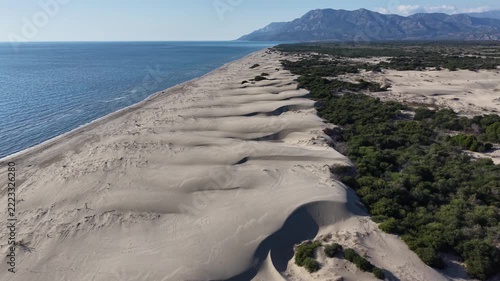 Aerial motion shot of long coastal sand dunes sliding beside calm blue sea with wind-sculpted ridges and sparse vegetation, dunes gradually shifting and curving inland toward dense green scrub and low