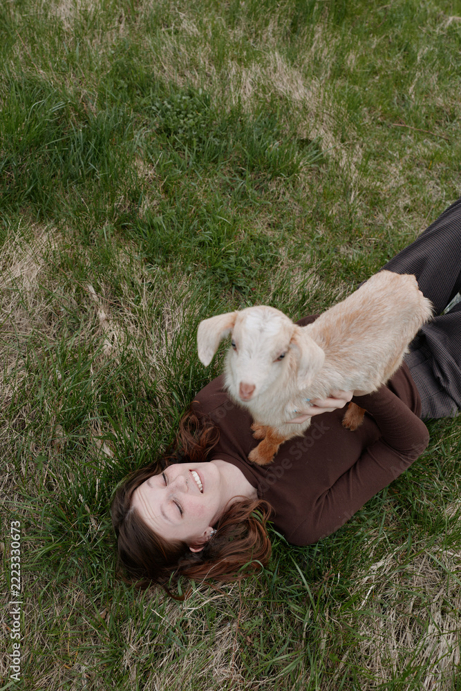 Fototapeta premium Relaxed woman lying on grass with a playful goat kid perched on her chest, outdoor lifestyle moment highlighting affection and cuddle, nature and companionship in a gentle, joyful scene.