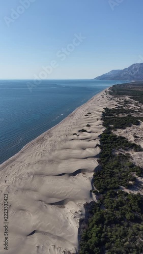Aerial view of long coastal sand dune field meeting calm blue sea under clear sky, wind-sculpted sand ridges and sparse vegetation lining shoreline with distant mountains on horizon, gentle midday sun
