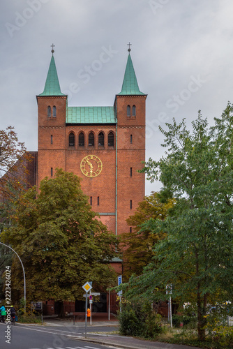 Fotografie Red-brick facade, twin copper-roofed spires, and prominent golden clock of the historic Erlöserkirche (Church of the Redeemer) framed by autumn trees in the Moabit district of Berlin, Germany