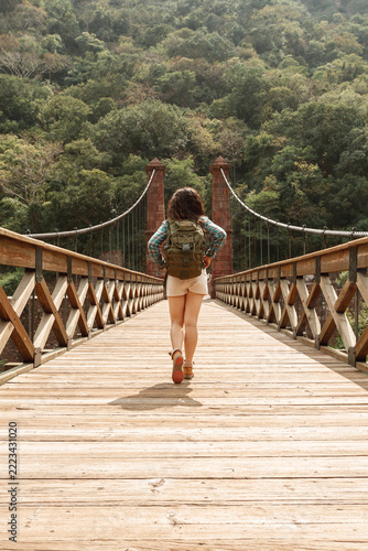 Rear View of Female Traveler with Backpack Walking Across Wooden Suspension Bridge