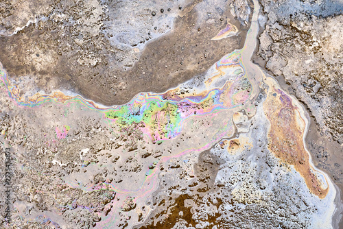 Iridescent gasoline layer covers muddy water puddle on street surface. Bright spectrum film shimmers over weathered asphalt ground, displaying hazardous petrochemical runoff after rainfall