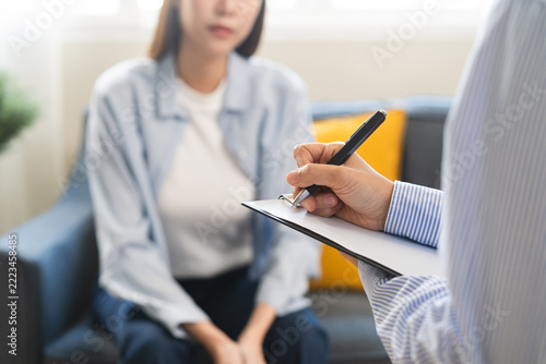 Female psychologist taking notes on clipboard during therapy session with a depressed Asian woman patient