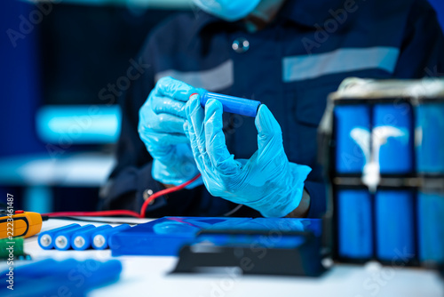 Close-up of a technician in blue gloves testing a cylindrical battery cell with probes in an electronics laboratory.