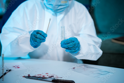 Forensic technician in protective clothing collects a sample with a swab and test tube at a crime scene laboratory table.