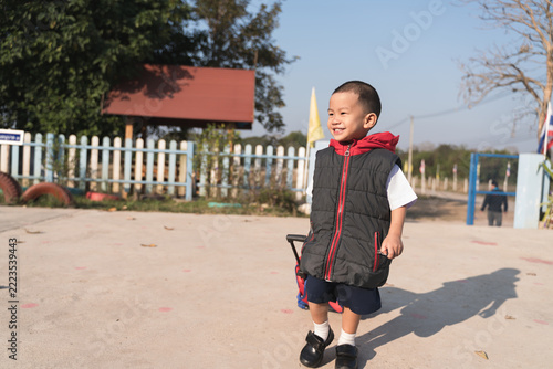 Happy young Thai boy walking to school in the morning sunlight, wearing a black and red jacket with a backpack trolley, smiling brightly outdoors.