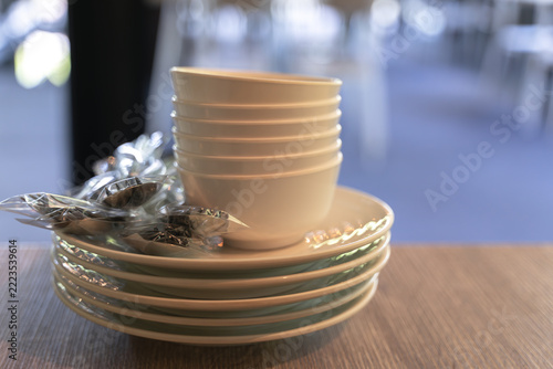 Stacked white bowls and plates with wrapped utensils on a wooden table in a dining area.