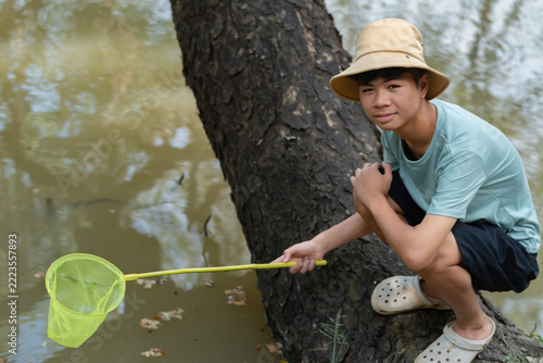 An Asian teenage boy sitting on a large tree trunk over a pond, holding a long yellow net to scoop water samples for biology field observation.