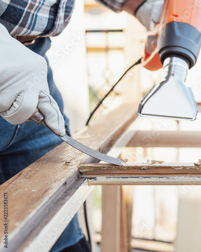 Adult carpenter wearing protective leather gloves, using a heat gun and a wood scraper to remove paint from an old wooden window. Construction industry, carpentry. Restoration. 