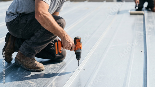 Roofer Kneeling on Metal Roof Using Orange Cordless Drill to Install Standing Seam Roofing Panel on Construction Site