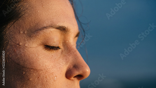 Close Up Profile of Woman Face with Closed Eyes and Sweat Droplets on Skin in Warm Golden Sunlight Against Blue Background with Copy Space