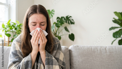 Sick Woman Blowing Nose into Tissue While Wrapped in Plaid Blanket on Sofa at Home with Cold and Flu Symptoms