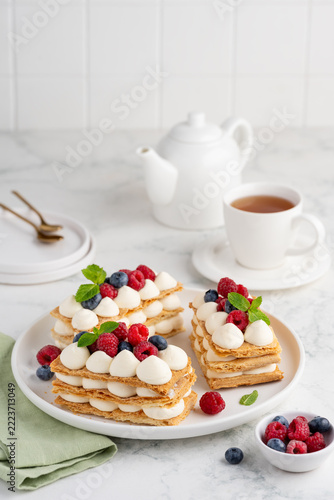 Delicious puff pastry Millefeuille desserts topped with fresh raspberries and blueberries, with a white teapot and cup in the background. Selective focus