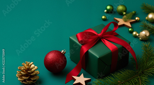    Modern flat lay on a teal background featuring a ruby-red bauble, gold pinecone, and forest-green gift with a crimson satin ribbon. Bold, contemporary holiday design.
