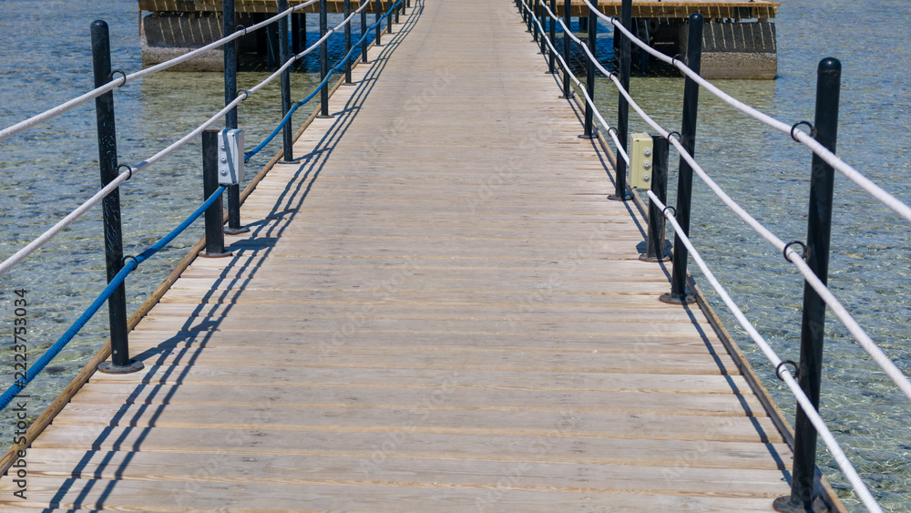 Naklejka premium Long wooden pier stretches over clear shallow tropical water with black metal railings and white rope barriers on both sides under bright sunny sky during day in coastal vacation setting.