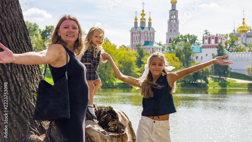 Smiling mother and her two daughters enjoy sunny day by lake with historic monastery in background.
