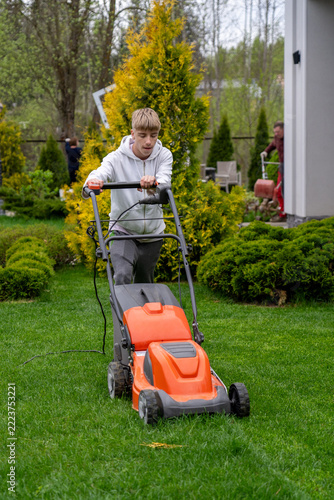 Young man wearing white hoodie mows green lawn in front of his house using orange electric lawn mower on spring afternoon while another person works in background.