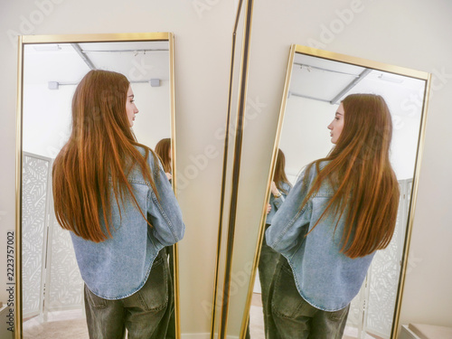 Young woman stands between two mirrors, facing away. Long brown hair flows down her back. She wears a denim shirt and dark jeans. Calm and confident, she checks her outfit. Perfect for fashion, beauty