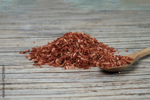Red rice grains spill from a wooden spoon. Rice rests on weathered, textured wood surface. Natural tones highlight wholesome, earthy quality. Ideal for health, nutrition, or culinary themes