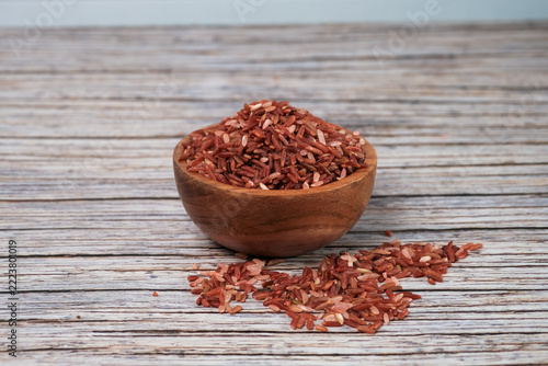 Red rice grains fill a rustic wooden bowl. Few grains spill out, resting on weathered wood. Natural light illuminates the texture and warm tones. Simple composition highlights organic