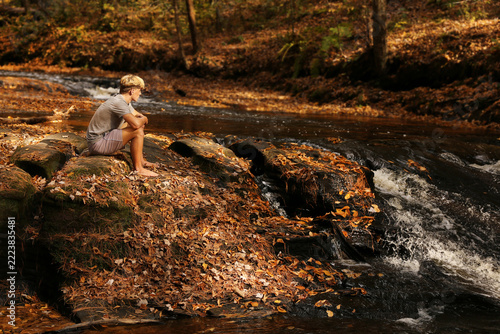 Teenage Boy Sitting on Rocky Waterfall on Fall Day Enjoying River and Forest