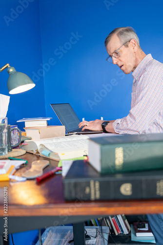 Elderly man focused on biblical translation work with open books and a laptop in home office.