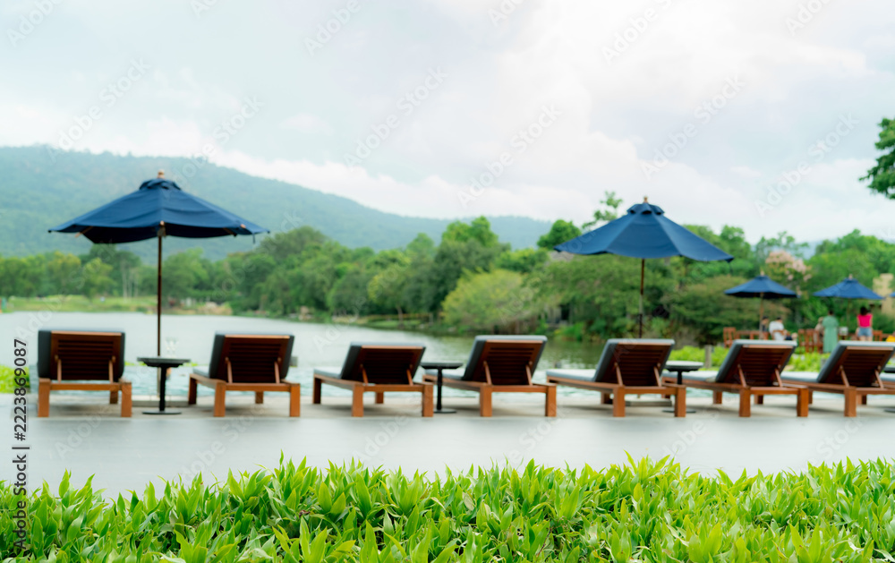 Fototapeta premium Luxury resort poolside with wooden lounge chairs and blue umbrellas beside a tranquil lake and green mountain during daytime, offering a tropical vacation retreat for relaxation and sustainable travel