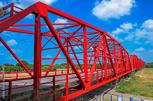 A striking red steel truss bridge stretching gracefully across the landscape, its vivid color contrasting with the serene greenery and bright sky.